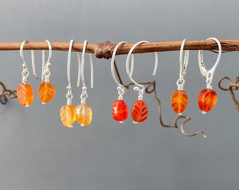 May include: A collection of six pairs of leaf-shaped earrings. The earrings are made of orange and yellow gemstones with silver hooks. The gemstones are carved with leaf details. The earrings are displayed on a brown twig against a gray background.