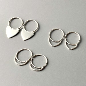 May include: Three pairs of silver earrings. One pair features a teardrop shape, while the other two pairs have a crescent design. The earrings are displayed on a gray surface.