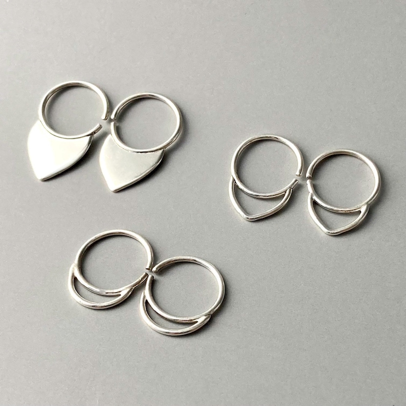 May include: Three pairs of silver earrings. One pair features a teardrop shape, while the other two pairs have a crescent design. The earrings are displayed on a gray surface.