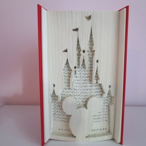 May include: A red book with the pages folded to create a 3D sculpture of Cinderella's castle from Disney World. The castle is white and has a Mickey Mouse head cut out in the front.