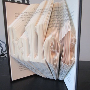 May include: A book with pages folded to spell out the word "HELLO". The letters are in white against the light-colored pages. The book has a black cover and is open, with the folded letters standing out.