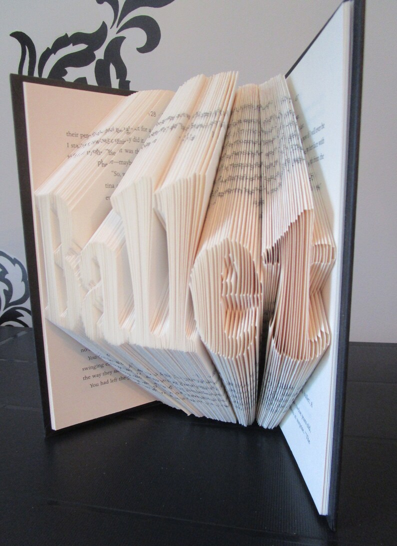 May include: A book with pages folded to spell out the word "HELLO". The letters are in white against the light-colored pages. The book has a black cover and is open, with the folded letters standing out.