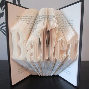 May include: A handmade book sculpture with the word "BEAUTY" folded into the pages. The book is open, revealing the sculpted letters in a three-dimensional design. The pages are cream-colored, and the book has a black cover.