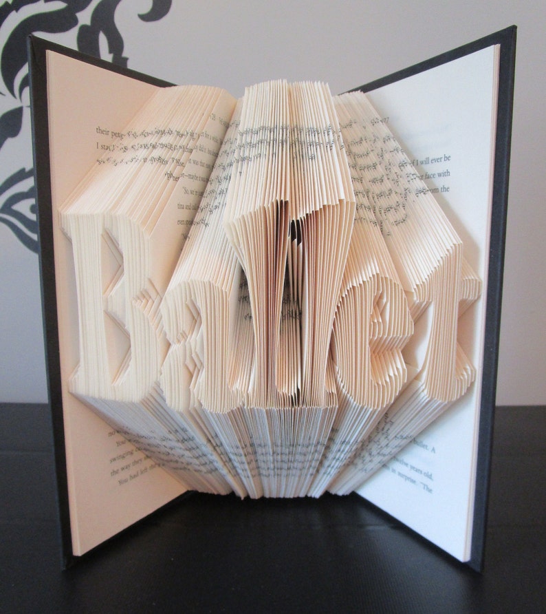 May include: A handmade book sculpture with the word "BEAUTY" folded into the pages. The book is open, revealing the sculpted letters in a three-dimensional design. The pages are cream-colored, and the book has a black cover.