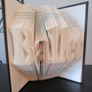 May include: A handmade book sculpture with the word "SECRET" folded into the pages. The book is open, revealing the sculpted letters in a three-dimensional design. The pages are a light beige color, and the book has a black cover.