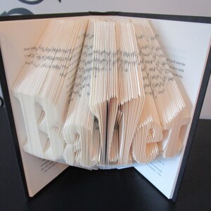 May include: A book with pages folded to spell the word "BEST". The book is open, with the folded letters prominently displayed in the center. The pages are cream-colored, and the book has a black cover.