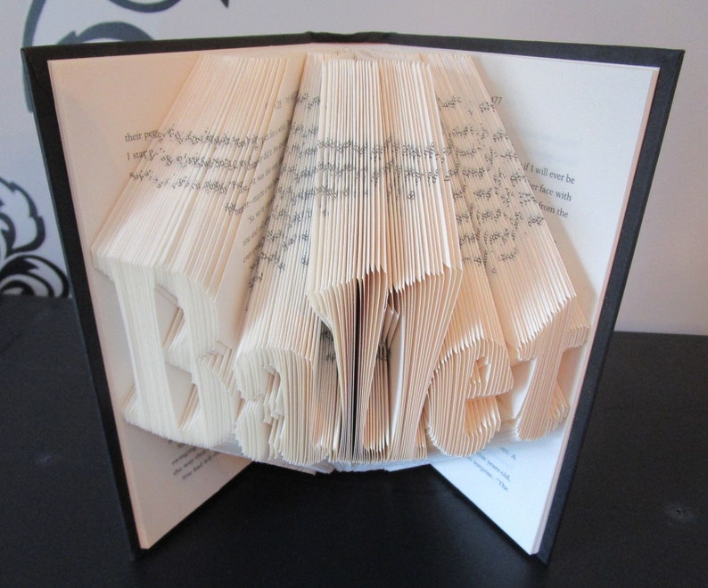 May include: A book with pages folded to spell the word "BEST". The book is open, with the folded letters prominently displayed in the center. The pages are cream-colored, and the book has a black cover.