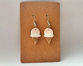 Puede incluir: Un par de pendientes colgantes con un diseño de cono de helado blanco y rosa. Los pendientes están hechos de arcilla polimérica y tienen ganchos plateados.