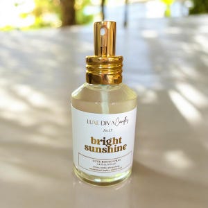 May include: A clear glass room spray bottle with a gold-colored top and a white label. The label reads "Luxe Diva Candles" and "bright sunshine" in gold lettering. The bottle contains a light yellow liquid.