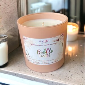 May include: A peach-colored candle in a glass jar with the text "Bubble Bath" on a label. The candle is hand-poured in Colorado and made with a coconut soy wax blend. The scent includes black currant, strawberry, raspberry, and vanilla.