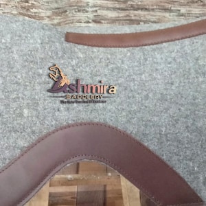 May include: A gray felt saddle pad with brown leather trim. The saddle pad has the text "Ashmira Saddlery" embroidered on it.