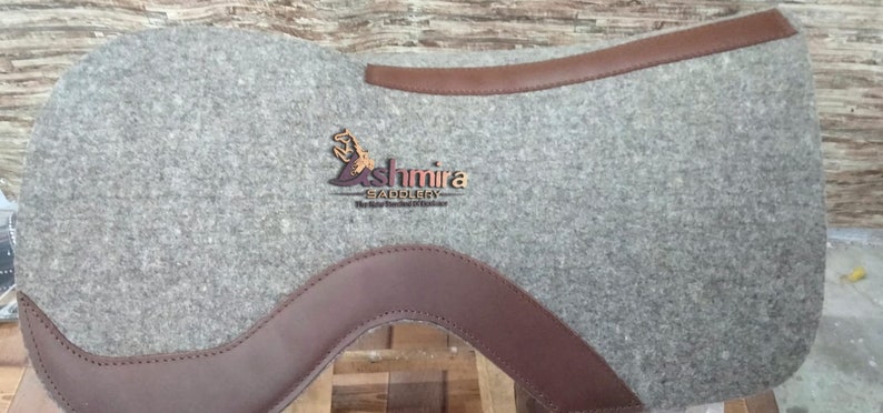 May include: A gray felt saddle pad with brown leather trim. The saddle pad has the text "Ashmira Saddlery" embroidered on it.