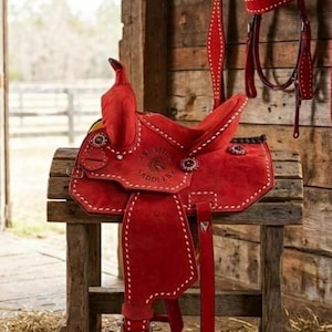 May include: A bright red suede western saddle featuring white contrast stitching, a horn, and a circular embossed logo reading Ashall Saddlery. A matching red bridle with white stitching hangs on the wall behind the saddle.