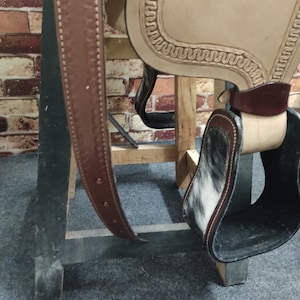 May include: A brown leather saddle with a white and black cowhide stirrup. The saddle is on a wooden stand.