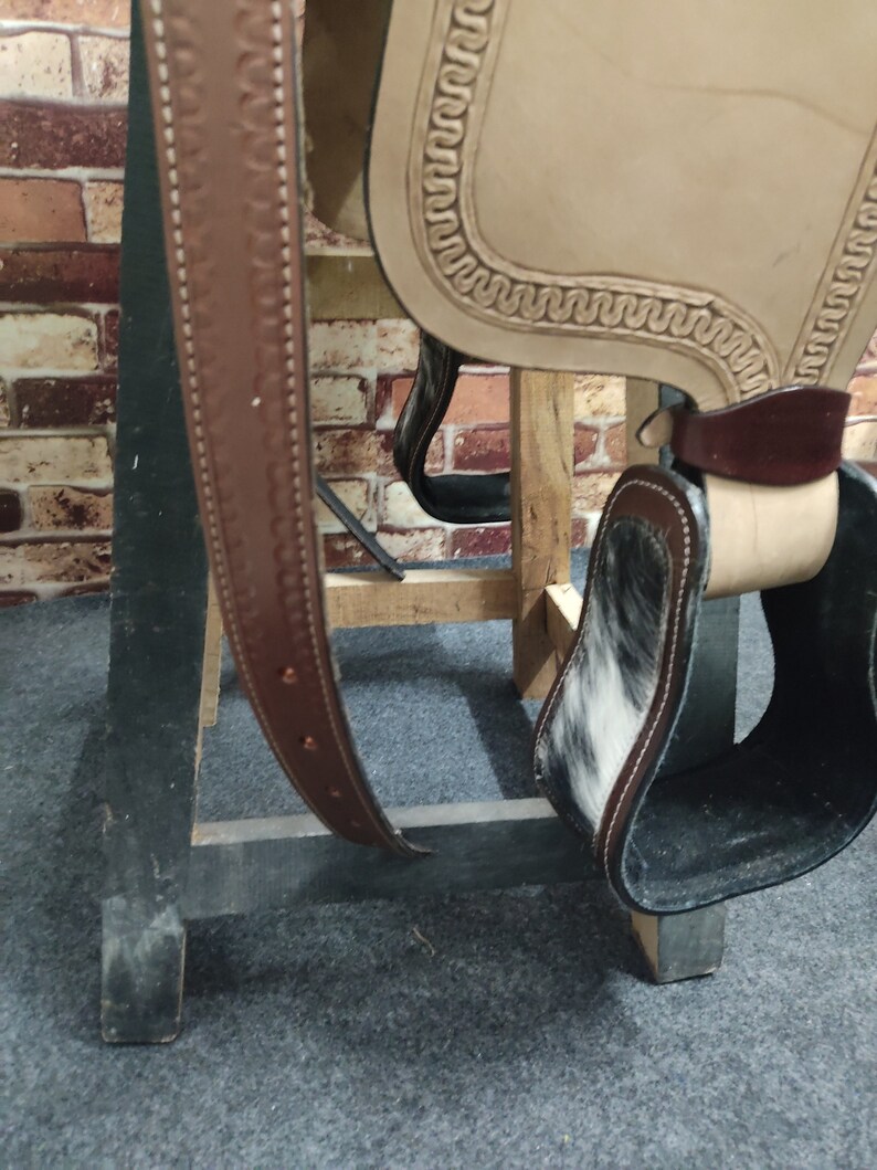 May include: A brown leather saddle with a white and black cowhide stirrup. The saddle is on a wooden stand.