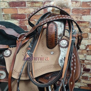May include: A close-up of a brown leather saddle with white stitching and a black leather strap. The saddle is decorated with silver conchos and has a white and brown cowhide seat. The saddle is against a brick wall background.