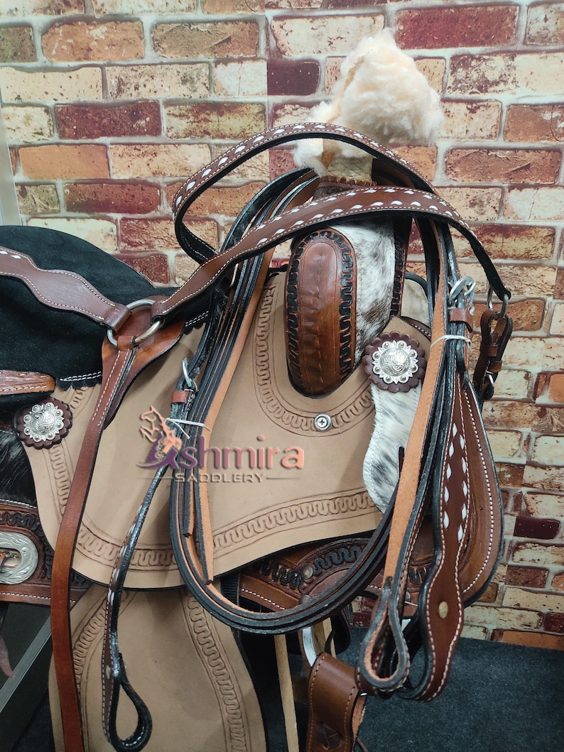 May include: A close-up of a brown leather saddle with white stitching and a black leather strap. The saddle is decorated with silver conchos and has a white and brown cowhide seat. The saddle is against a brick wall background.
