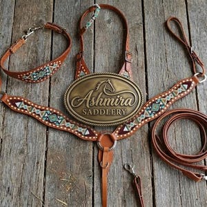 May include: A brown leather horse breast collar and bridle set featuring intricate geometric beadwork in turquoise, white, and black. A large oval brass plate in the center is embossed with the text Ashmira Saddlery and a horse head silhouette.