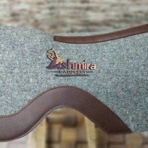May include: A gray felt saddle pad with brown leather trim. The pad features the logo "Ashmira Saddlery" in brown embroidery.
