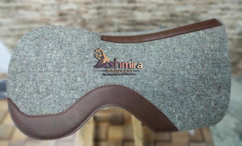 May include: A gray felt saddle pad with brown leather trim. The pad features the logo "Ashmira Saddlery" in brown embroidery.