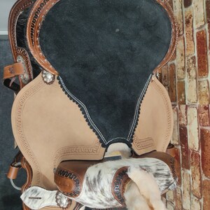 May include: A brown leather saddle with a black suede seat and white and brown hair accents. The saddle features a decorative stitching pattern and silver conchos.