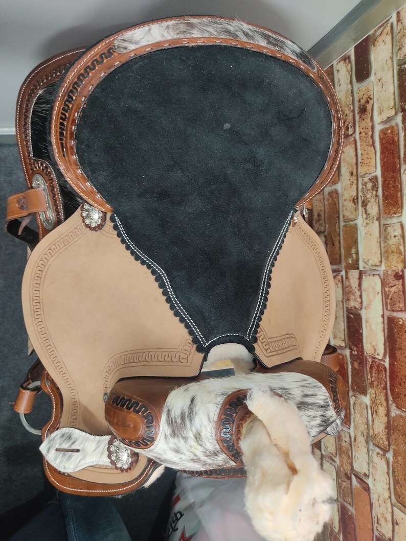 May include: A brown leather saddle with a black suede seat and white and brown hair accents. The saddle features a decorative stitching pattern and silver conchos.