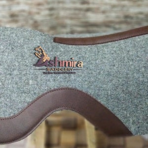 May include: A gray felt saddle pad with brown leather trim. The pad features the logo "Ashmira Saddlery" in a brown and gold color scheme.