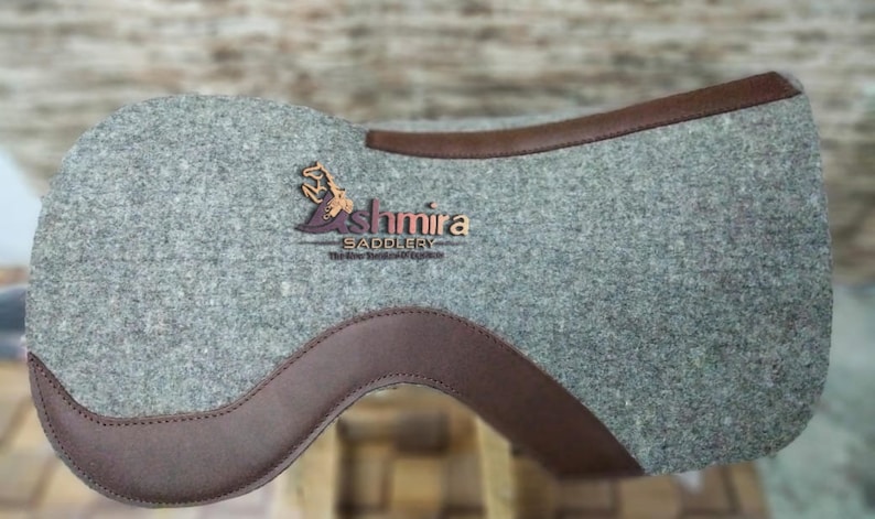 May include: A gray felt saddle pad with brown leather trim. The pad features the logo "Ashmira Saddlery" in a brown and gold color scheme.