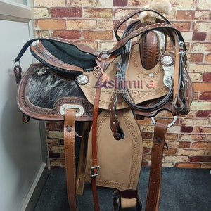 May include: A brown leather saddle with a black and white cowhide seat. The saddle has silver accents and is decorated with stitching. The saddle is on a wooden stand and has a leather stirrup.