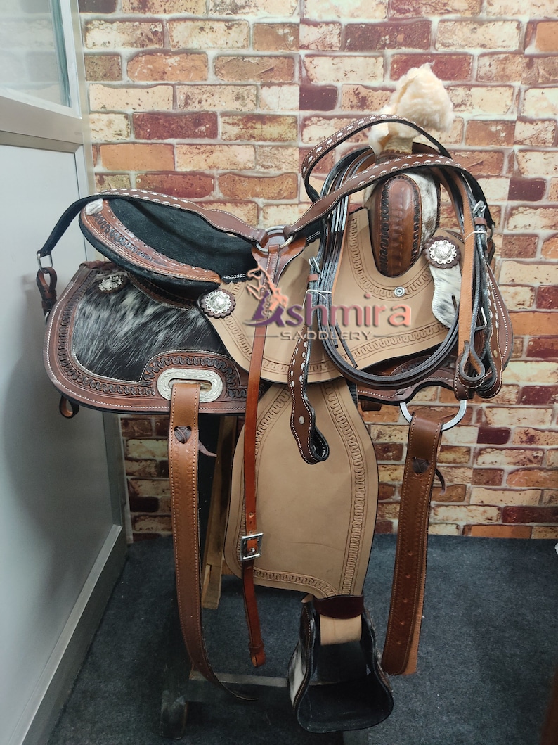 May include: A brown leather saddle with a black and white cowhide seat. The saddle has silver accents and is decorated with stitching. The saddle is on a wooden stand and has a leather stirrup.