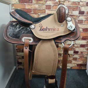 May include: A brown leather saddle with a black seat and a white and black cowhide pattern. The saddle has a silver concho and the text "Ashmira Saddlery" is embossed on the saddle.