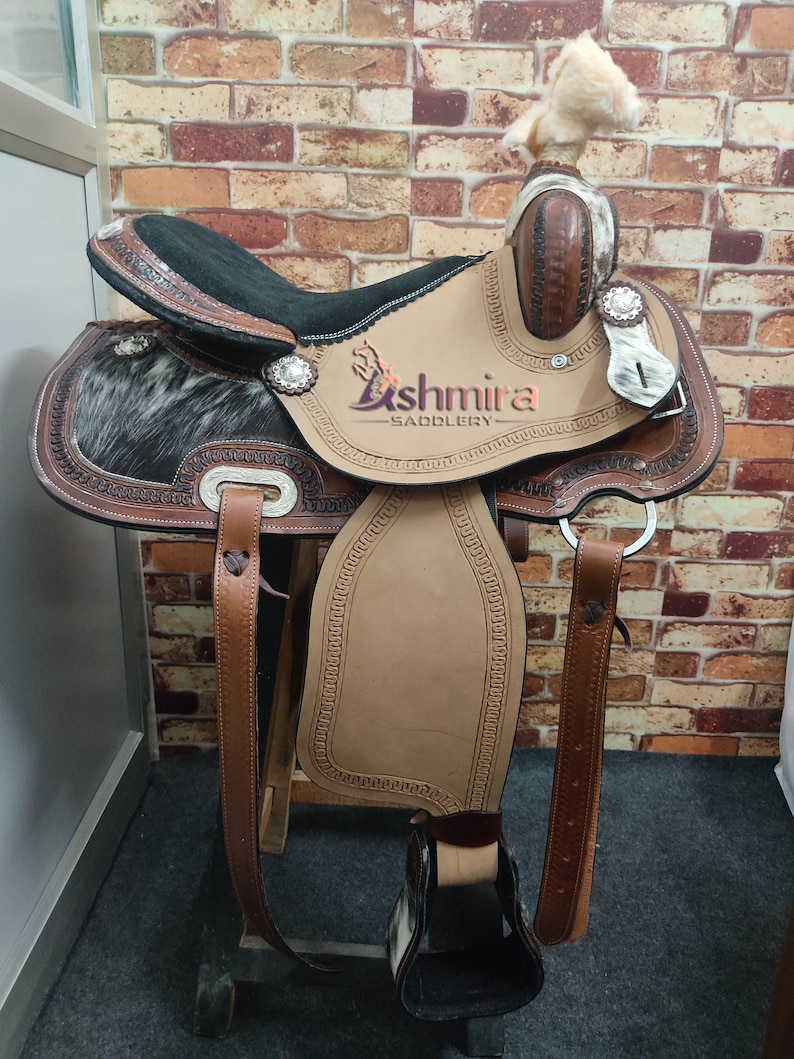 May include: A brown leather saddle with a black seat and a white and black cowhide pattern. The saddle has a silver concho and the text "Ashmira Saddlery" is embossed on the saddle.