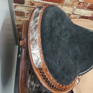 May include: Close-up of a black leather saddle seat with brown leather trim and a white and black cowhide pattern. The saddle seat is decorated with intricate stitching.