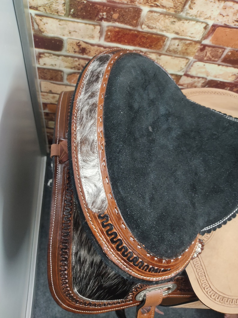 May include: Close-up of a black leather saddle seat with brown leather trim and a white and black cowhide pattern. The saddle seat is decorated with intricate stitching.
