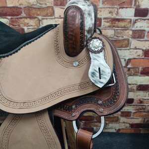 May include: A brown leather saddle with a white and brown patterned design. The saddle has a silver concho and a leather stirrup.