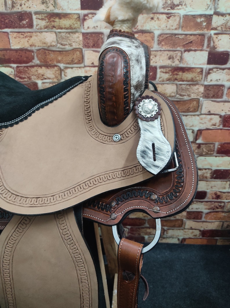 May include: A brown leather saddle with a white and brown patterned design. The saddle has a silver concho and a leather stirrup.