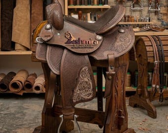 May include: A brown leather western saddle featuring intricate floral tooling and basket-weave patterns. The saddle displays a prominent logo with the text "Kashmira Saddlery" on the side, silver-toned conchos, and a metal stirrup, mounted on a wooden stand.