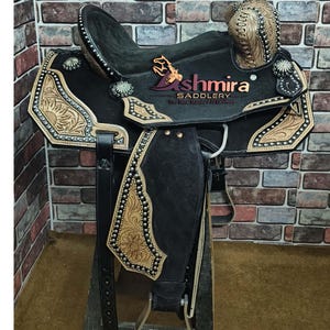 May include: A black leather saddle with intricate floral tooling and silver accents. The saddle is on a wooden stand and has a  "Ashmira Saddlery" logo with the tagline "The New Standard of Excellence".