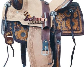 May include: A western saddle featuring dark brown leather with intricate floral tooling and sunflower patterns, contrasted by a light tan suede seat and fenders. The saddle includes silver-toned hardware, a horn, and the Ashmira Saddlery logo.