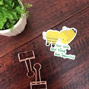 May include: Two gold binder clips and a sticker with two cartoon bees and the text "Bee safe, Bee kind, Bee impactful".