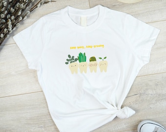 May include: White t-shirt with a yellow graphic of teeth with plants growing out of them. The text "Keep going, keep growing" is above the teeth.
