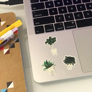Puede incluir: Tres pegatinas de dientes con plantas verdes y negras que les salen, colocadas en un port&aacute;til plateado.