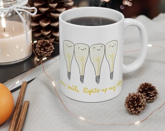Puede incluir: Taza de cerámica blanca con asa negra. La taza tiene un diseño de cuatro bombillas con caras. El texto "your smile lights up my day" está impreso en la taza.