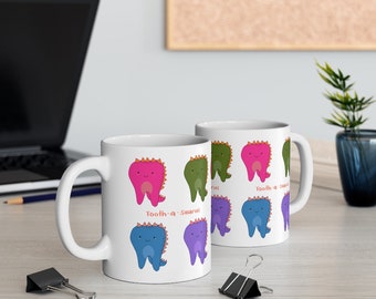 Puede incluir: Dos tazas de cerámica blanca con un diseño de dientes de dibujos animados coloridos con características de dinosaurio. El texto "Tooth-a-saurus" está impreso en las tazas.