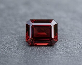 May include: A faceted, rectangular-cut, deep red gemstone. The gem has a rich, dark color and is set against a gray, textured background. The cut of the gem creates multiple facets that reflect light.