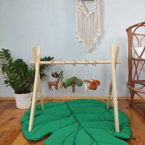 May include: Wooden play gym with a green leaf-shaped play mat. The gym has five hanging toys, including a deer, a tree, a mushroom, and a squirrel.