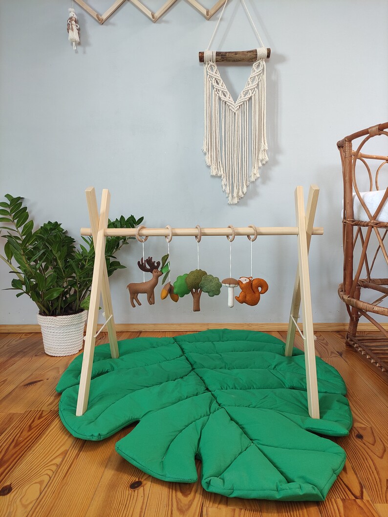 May include: Wooden play gym with a green leaf-shaped play mat. The gym has five hanging toys, including a deer, a tree, a mushroom, and a squirrel.