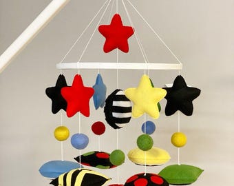 Puede incluir: Móvil de bebé colorido con estrellas de fieltro, círculos y otras formas en rojo, negro, amarillo, azul y verde. El móvil está suspendido de un marco blanco e incluye varias formas y tamaños.
