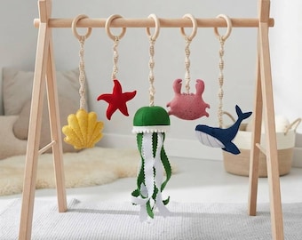 May include: A wooden baby play gym frame with five hanging felt sea creatures including a yellow seashell, a red starfish, a green jellyfish with white ribbon tentacles, a pink crab, and a blue whale, all suspended by macrame cords from wooden rings.