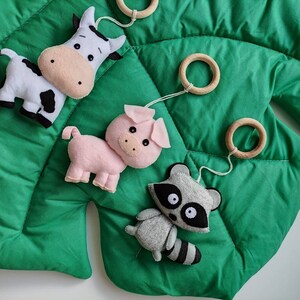 May include: A green felt play mat with a cow, pig, and raccoon hanging from wooden rings. The animals are made of felt and have embroidered details.